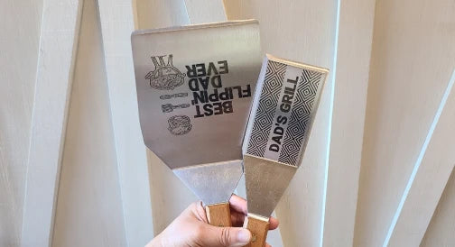 Two personalized grill spatulas with engraved designs and text held in hand.